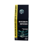 A 12oz box of Broken Road Coffee Company Bourbon Minerio Dark Roast coffee, featuring a dark navy blue and black design with a bright yellow vertical banner on the left and a scenic mountain graphic.