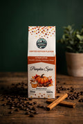 A package of South Road Coffee Company Pumpkin Spice coffee roast, standing on a rustic wooden table scattered with coffee beans and cinnamon sticks, with a dark green background and a blurred potted plant.