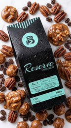 A black coffee bag labeled 'Reserve Collection' with mint green accents, surrounded by roasted coffee beans, pecans, and caramel-coated nuts on a white background.
