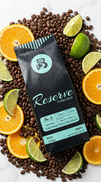 A bag of Reserve Collection No. 3 coffee lies on a white marble surface surrounded by scattered coffee beans, sliced oranges, and lime wedges.